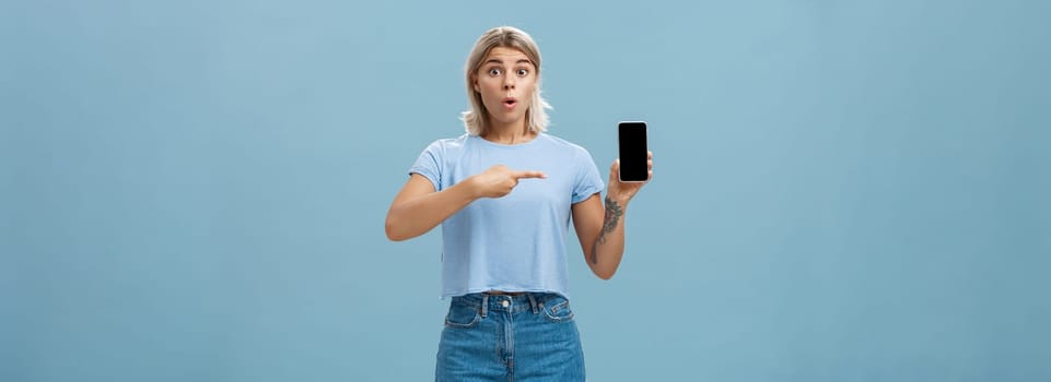 Wow look what found online. Portrait of impressed speechless good-looking young sportswoman in t-shirt and denim shorts saying wow folding lips showing smartphone and pointing at screen over blue wall. Technology concept