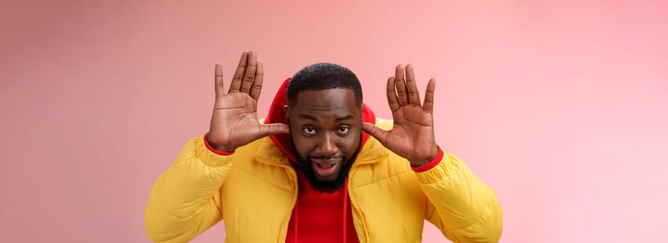 Ha cannot hear you sorry. Portrait ignorant funny careless unbothered young african guy pretending dumb plug thumbs ears show palms bending camera eavesdrop, standing pink background.