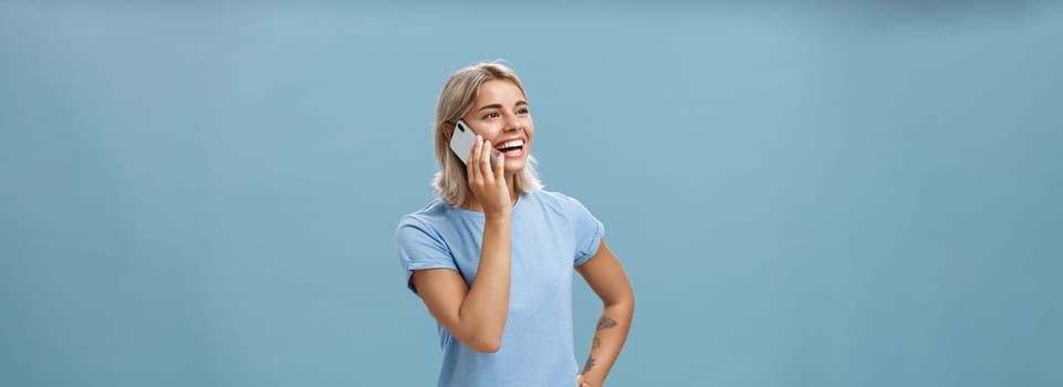 Lifestyle. Waist-up shot of sociable amused and happy attractive caucasian fair-haired woman in casual t-shirt standing half-turned gazing left with hand on hip while talking on smartphone over blue background.