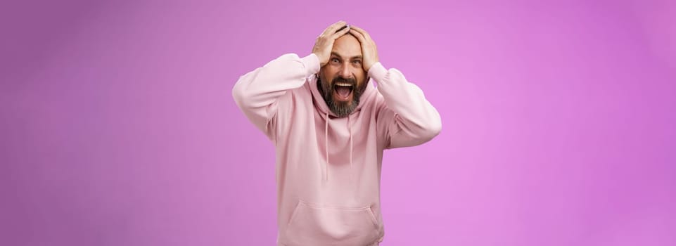 Outraged furious mad adult man bearded yelling shocked despair hatefully looking camera insane holding hands head frowning shouting losing bet disappointed very upset standing purple background. Lifestyle.