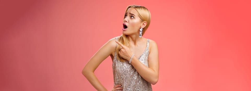 Shocked displeased bothered arrogant blond glamour woman in silver glittering dress turning upper right corner pointing complaining strange noise come upstairs, standing questioned red background.