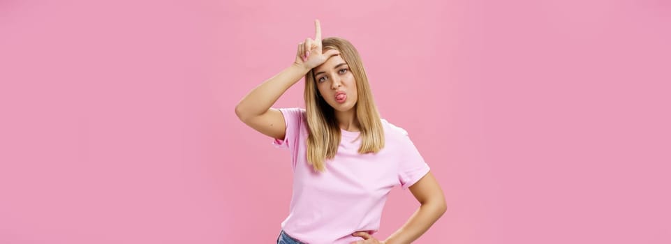 Girl mocking under boyfriend showing loser sign on forehead sticking out tongue gazing arrogant and indifferent at camera being moody holding hand on waist posing against pink background. Body language concept