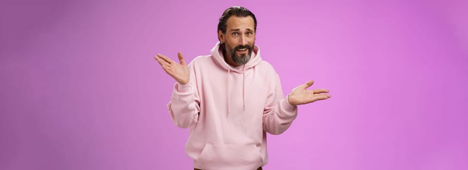 So what bite me. Portrait ignorant careless cool stylish mature bearded man earring pink hoodie shrugging hands sideways mocking being rude standing pissed unwilling help standing purple background.