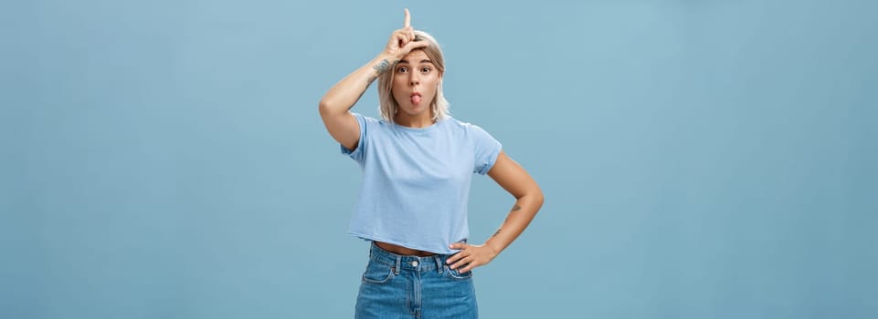 Lifestyle. Studio shot of playful triumphing good-looking blonde female with tattoed arms showing letter L on forehead and sticking out tongue mocking over loser and celebrating own victory over blue wall.