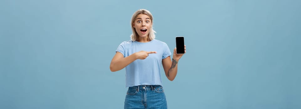 Studio shot of amazed and impressed charming european blonde in casual clothes gasping charmed at camera while showing smartphone screen pointing at device with finger over blue background. Technology and advertisement concept