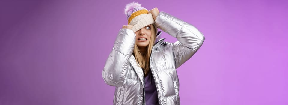 Awkward embarrassed cute blond girl worried pulling hat face hiding from ex-boyfriend pretend not see him standing anxiously clenching teeth peeking one eye left nervous, purple background.