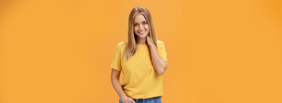 Portrait of shy and timid feminine girl with tan and straight fait hair rubbing neck and smiling sensually with happy carefree expression holding hand in pocket posing against orange background. Lifestyle.