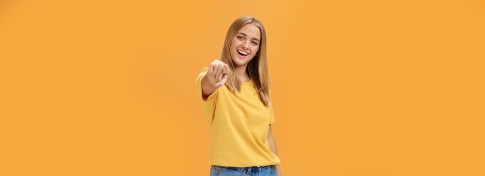 Woman directing at camera with index finger and smiling friendly as if talking to us standing amused and happy with confident carefree expression pulling arm towards to point over orange background. Lifestyle.
