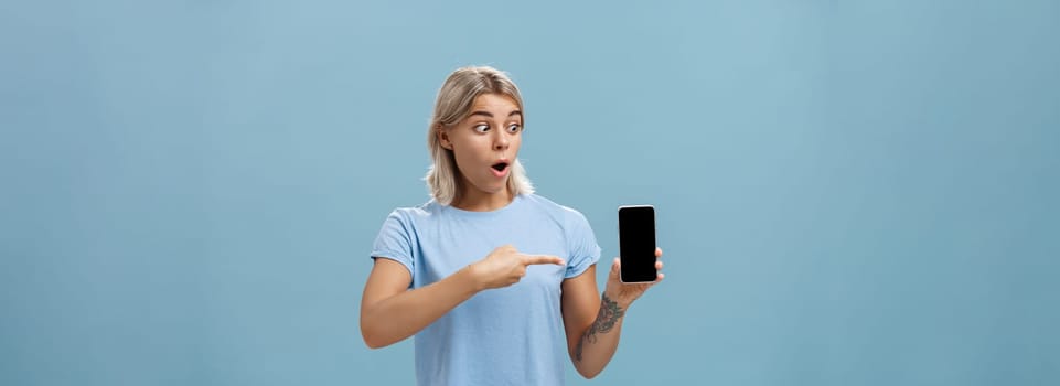 Girl being amazed with cool new smartphone received on birthday. Surprised speechless happy woman with fair hair and tattoo dropping jaw holding smartphone pointing and staring at gadget screen.