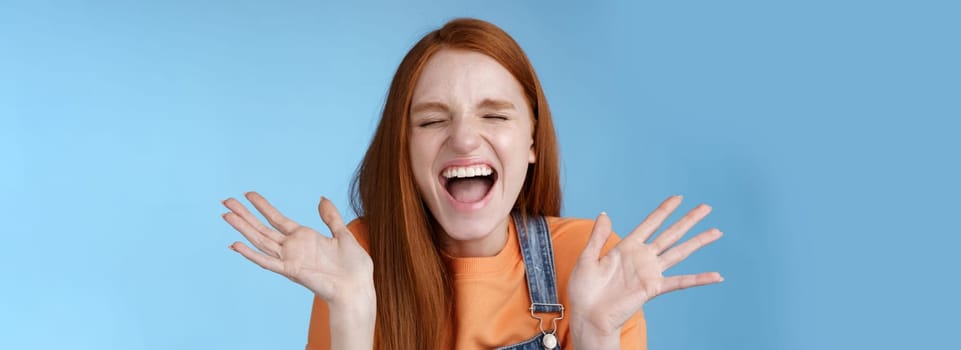 Positive carefree triumphing young attractive redhead girl scream out loud joy happiness close eyes waving raised hands win first prize dream come true celebrating victory good news, blue background.