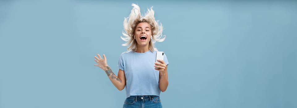 Expressing happiness with help of great tunes. Joyful amused and happy good-looking young female student jumping having fun listenign music in wireless earbuds, holding smartphone over blue wall.