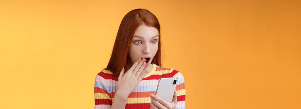 Lifestyle. Amazed speechless young teenage redhead girl student gasping drop jaw say omg wow cover opened mouth palm look shocked surprised smartphone display reading fresh gossips orange background.