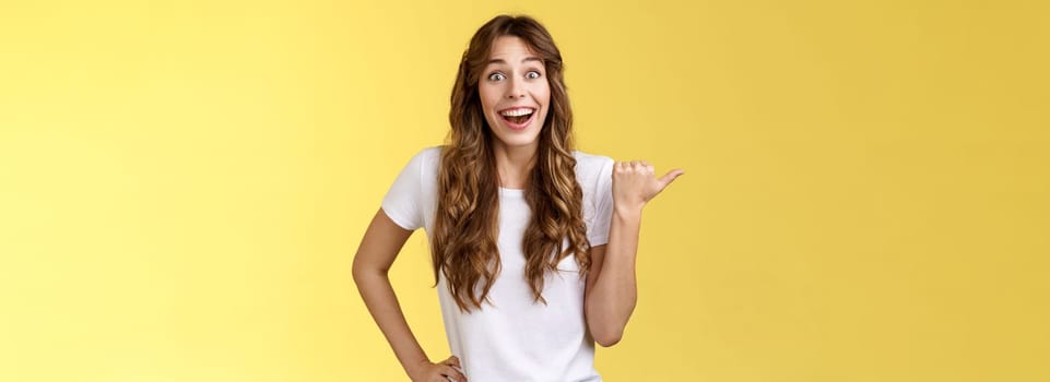 So amazing lets go together. Lively excited european female curly haircut wanna visit cool party smiling joyfully pointing thumb left grinning thrilled look camera admiration yellow background.