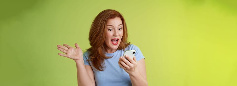 Surprised glad middle-aged pretty redhead woman look smartphone raise hand celebratory excitement open mouth. reacting impressed gladly read good excellent news look mobile screen green background.