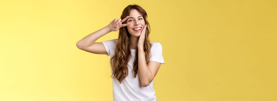 Cheerful positive lovely girlfriend curly hairstyle touch cheek blushing modest cute showing victory peace sign near eye optimistic upbeat attitude having fun enjoying summer yellow background. Lifestyle.
