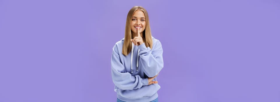 Portrait of joyful cute feminine young woman in hoodie smiling happily showing shush gesture hiding surprise asking keep secret standing amused and carefree against purple background. Copy space