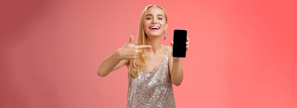 Boastful proud charming elegant blond woman in stylish evening dress show smartphone display proudly pointing mobile phone screen smiling showing photo boyfriend, standing red background.
