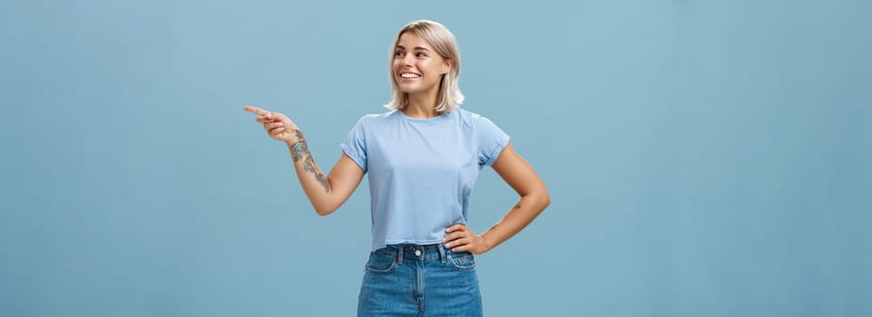Bossy confident female manager with tattoo on arm holding hand on waist pointing and gazing right with pleased relaxed look giving directions to employees posing over blue background. Copy space