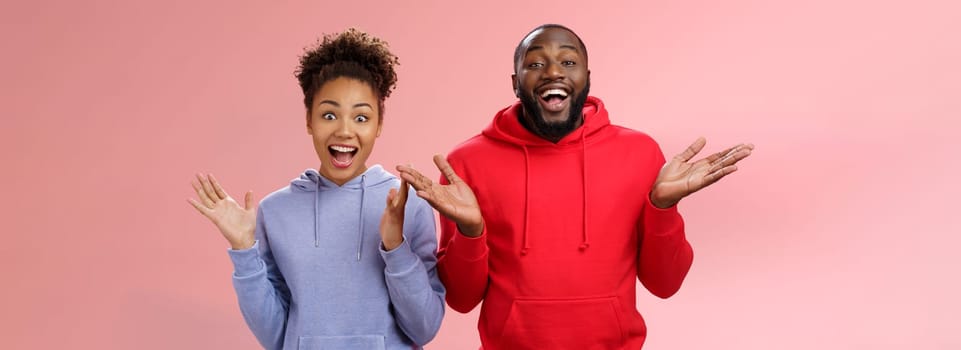 Guys receive nice surprise. Two attractive carefree surprised happy african man woman clapping hands joyfully greeting welcoming guests inviting people joing party standing friendly pink background.