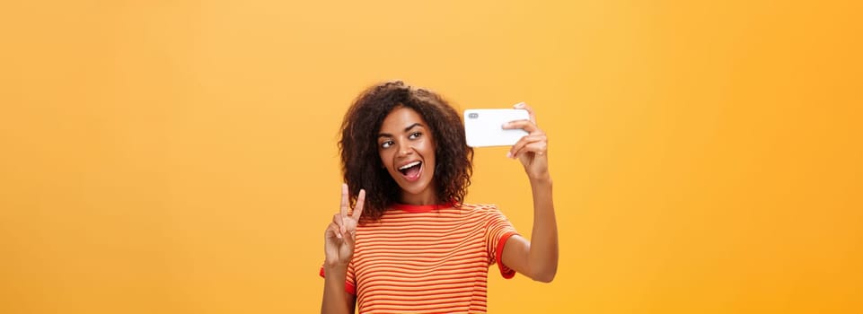 Smile to camera. Attractive and stylish self-assured dark-skinned female model with curly hairstyle showing peace gesture while taking selfie holding smartphone near face and smiling at gadget screen.