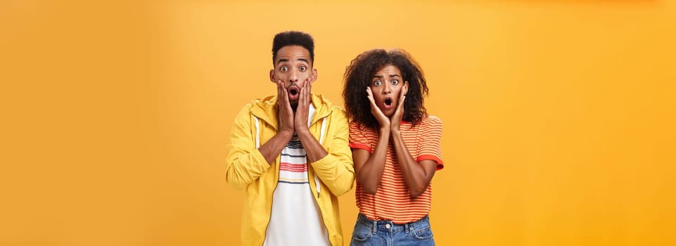 Portrait of shocked and stunned speechless girlfriend dropping jaw from amazement with boyfriend feeling amazed from shook news posing together surprised and astonished over orange wall. Lifestyle.