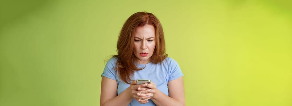 Lifestyle Concept - Confused unsure redhead middle-aged woman learn how use social media. trying understand emoji look intense focused smartphone display reading important news message stand green background.
