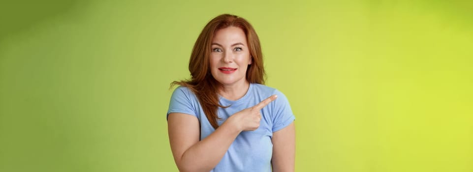 Surprised excited middle-aged wondered redhead woman pointing left amused standing thrilled joyful green background look camera curious interested cannot wait check-out great promo.