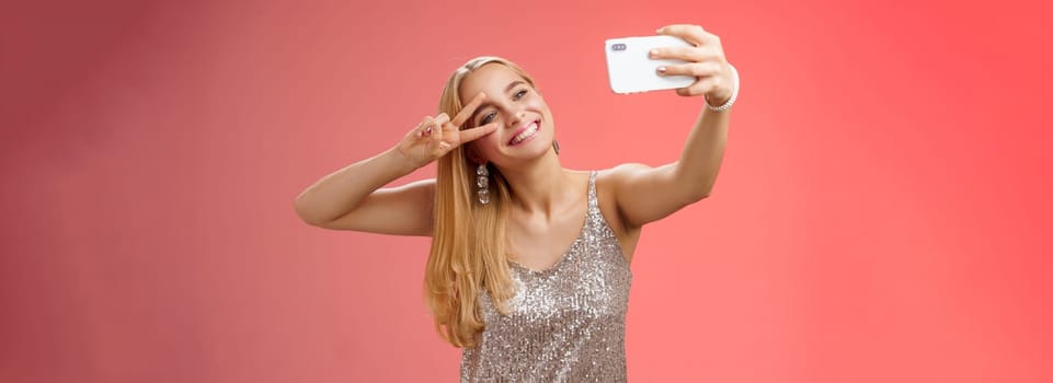 Lifestyle. Stylish fabulous glamour young blond woman in glittering silver dress tilting head carefree show peace gesture victory sign extend arm holding smartphone taking selfie recording video post online.