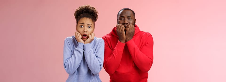 Shocked scared two insecure young african american couple both frightened spiders standing bed terrified freeze from fear holding hands mouth biting fingers shaking horror, pink background.