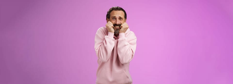 Scared insecure silly adult bearded man grey hair in pink hoodie press palms mouth bite fingers clench teeth shocked frightened widen eyes terrified standing stupor horrified, purple background.