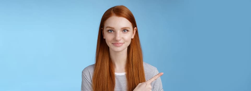 Attractive modest good-looking redhead girlfriend acting mature friendly smiling pointing left index finger giving direction show way bathroom standing blue background joyful kind grin.