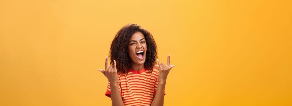 Waist-up shot of amazed happy stylish african american woman feeling awesome rocking on party yelling from joy and satisfaction showing rock n roll gesture posing over orange background. Copy space