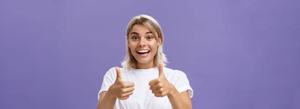 Lifestyle. Waist-up shot of optimistic and supportive good-looking caucasian female in white t-shirt cheering and smiling broadly hearing awesome idea approving it with thumbs up gesture over purple wall.