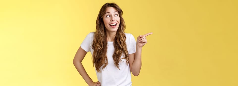 Wondered impressed charismatic fascinated smiling happy girl pointing look upper left corner speechless surprised grinning toothy happiness joy expression contemplate great view yellow background.