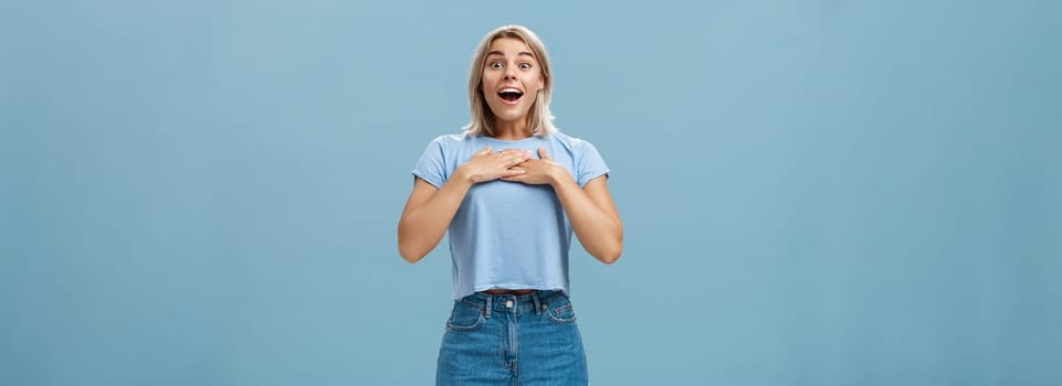 Lifestyle. Portrait of amazed and charmed attractive blond female student with tanned skin in denim shorts and summer t-shirt holding palms on breast gasping and smiling joyfully being grateful and pleased.