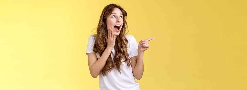 Silly flirty young attractive woman sighing admiration excitement checking out amazing stunning promo drop jaw smiling touch cheek touched delighted turn pointing left amused yellow background. Lifestyle.