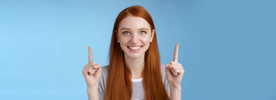 Amused excited happy smiling redhead girl look pointing up grinning gladly watching awesome open air perfomance standing joyfully showing cool thing indicating product, blue background.