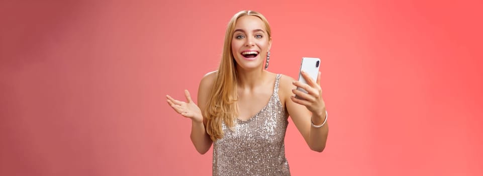 Astonished happy charming blond girl in silver glittering stylish dress holding smartphone amazed liking awesome result edit photo app smiling wondered amused, standing red background.