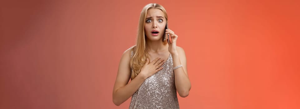 Lifestyle. Concerned worried woman blond hairstyle in silver stylish glittering dress hold hand heart widen eyes afraid gasping speechless listen terrifying news talking smartphone express empathy worry.