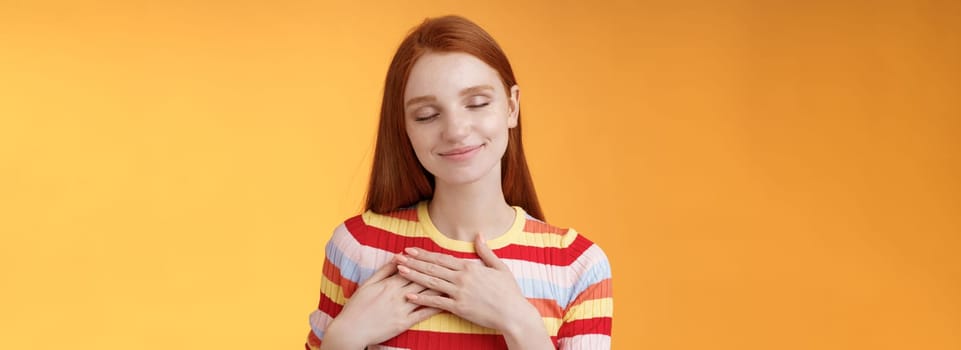 Heart keeps warm save dearest memories inside. Smiling tender heartwarming redhead lovely girl touch chest grinning closed eyes recall lovely nice moment standing romantic orange background.