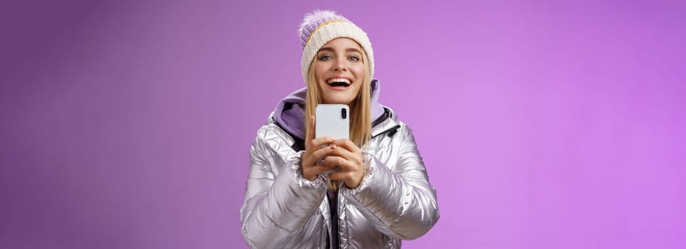 Amused excited attractive blond girlfriend holding smartphone up recording video boyfriend step snowboard first time capturing memories mobile camera standing happily purple background.