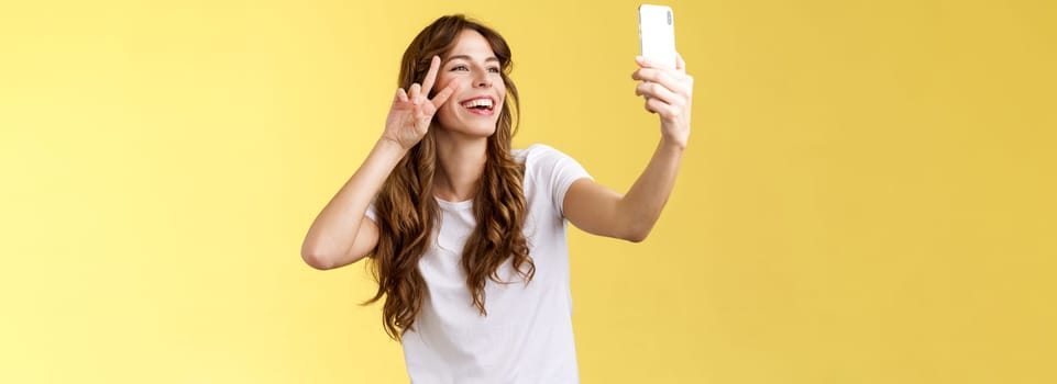 Positive charisamtic happy young girl curly-haired raise smartphone taking selfie show peace victory sign having video call stand yellow background smiling mobile front camera posing lively. Lifestyle.