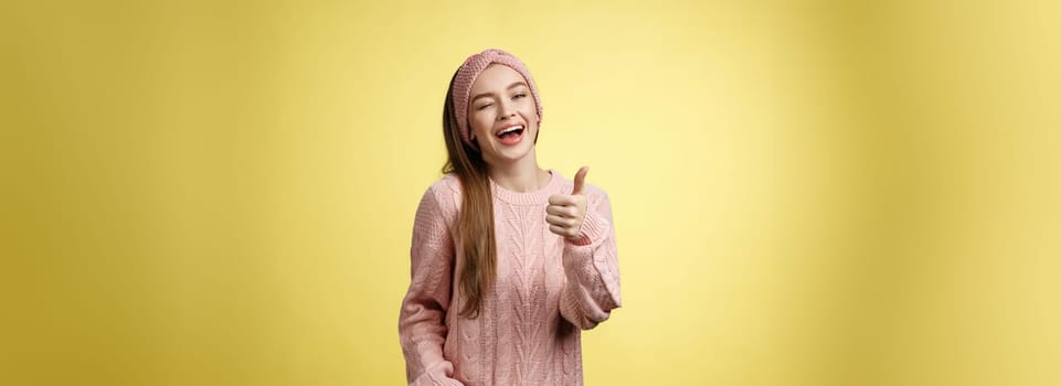 Happy amused beautiful young european girl in sweater, knitted headband showing good gesture, satisfied with service provided, winking sassy smiling with thumb-up in approval, giving recommendation.
