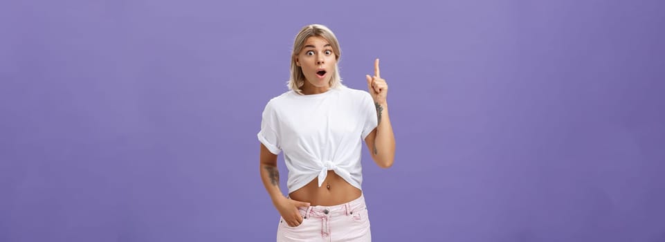 I have got idea. Thrilled and excited attractive stylish feminine woman with tattoos popping eyes folding lips, gasping raising index finger up in eureka gesture having excellent idea over purple wall.