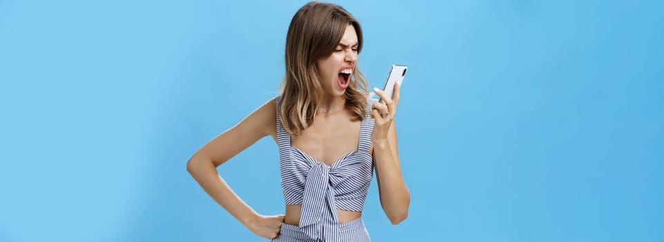 Woman yelling at smartphone with sick and tired look being agressive and angry swearing on boyfriend who dumbed her on phone holding hand on waist looking with hate at cellphone screen. Technology and emotions concept