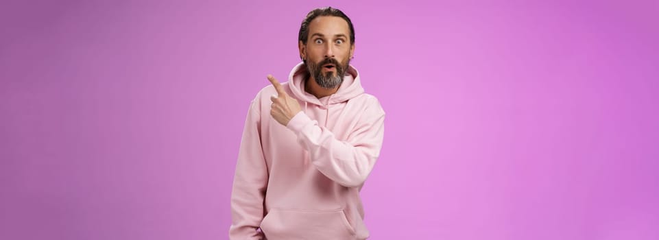 Amused handsome hipster mature 40s bearded caucasian man grey hair wearing trendy pink hoodie fold lips amazed wow pointing upper right corner widen eyes surprised check out awesome product. Lifestyle.