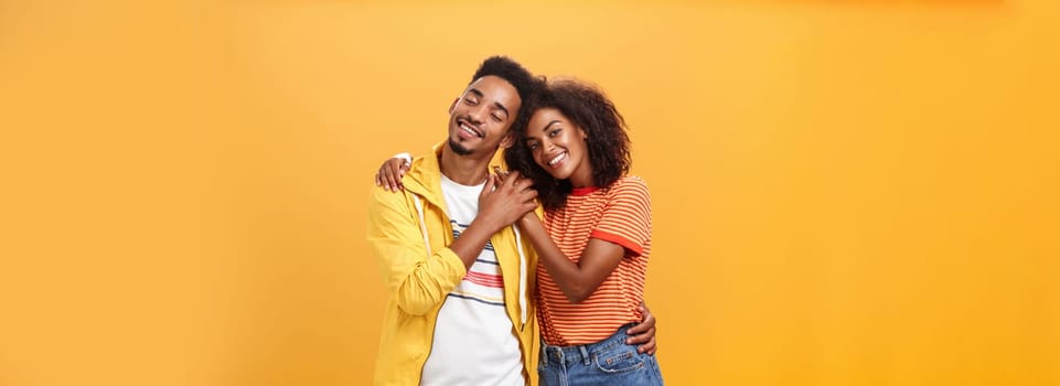 They love each other. Portrait of two charming african american man and woman in relationship hugging with heartwarming smile touching hands smiling gently posing against orange background.
