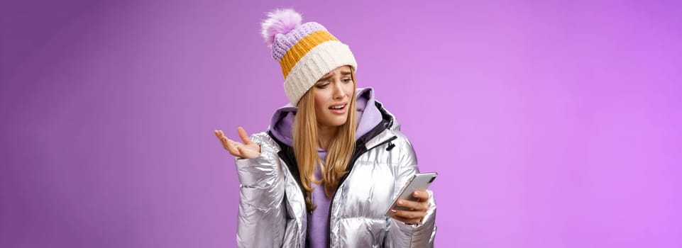 Upset disappointed attractive whining gloomy blond girl in silver jacket standing outside hat holding smartphone shrugging raising hand dismay complaining slow mobile internet, purple background.