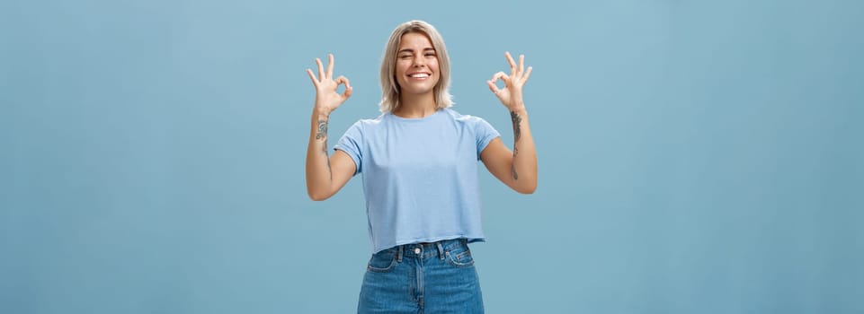 I got you covered. Enthusiastic attractive and energized female blonde with tattooed arms showing okay or perfect sign and winking to assure friend everything done smiling joyfully over blue wall.
