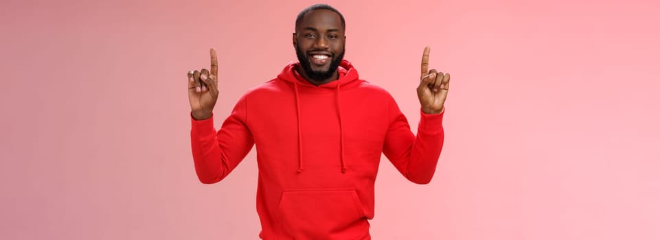 Devious cute african-american bearded guy in red hoodie smiling mysterious know exactly what you want pointing raised index fingers up grinning show perfect copy space promo, pink background.